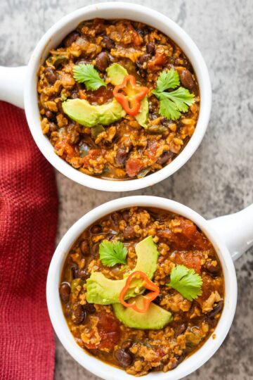 Two bowls of TVP black bean chili with avocado on a red cloth.