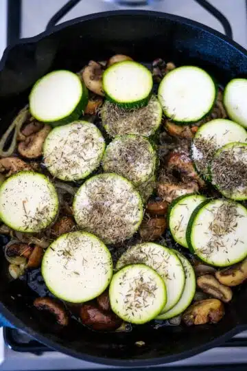 Zucchini and mushrooms sizzling in a skillet on a stove.