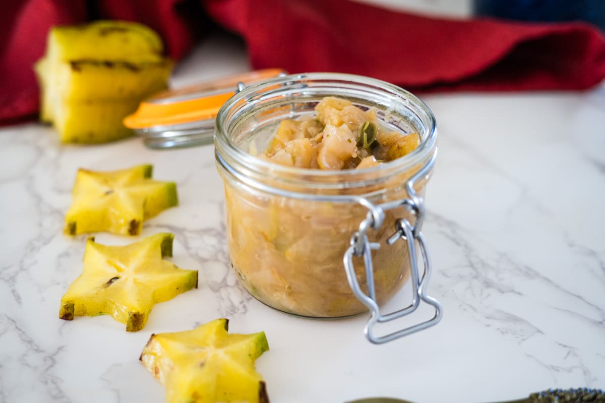 A jar with pineapple slices and a spoon next to it, filled with star fruit chutney.